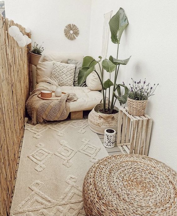 a cute boho balcony done in creamy shades, with a low seat with boho pillows, a beautiful rug, a crate and a basket with a plant, a jute pouf and some candle lanterns