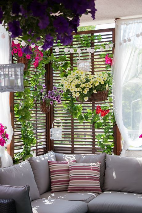 a balcony with a corner sofa and pillows, a trellis with blooms and greenery, some lights and a curtain