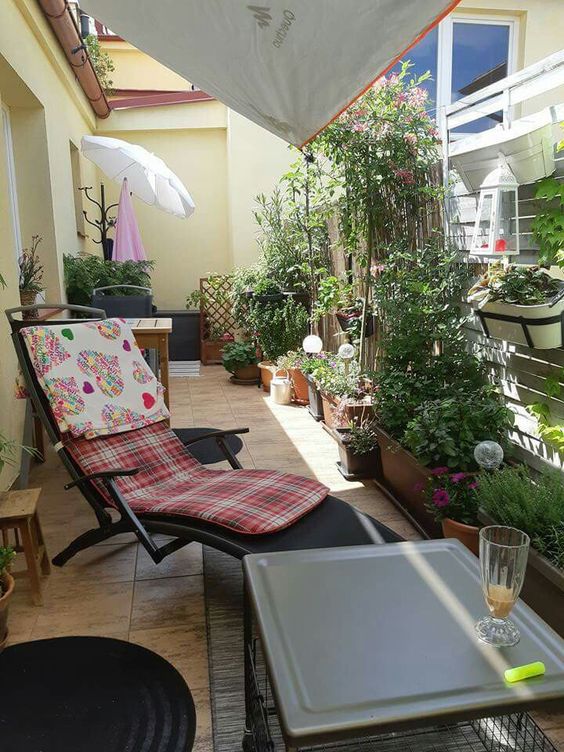 a balcony with greenery planters, blooms and a trellis with greenery, a lounger with cushions, side tables and stools