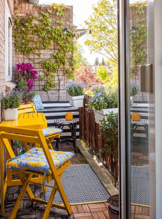 a balcony with potted blooms and greenery, bold yellow furniture, a trellis on the wall and some vines climbing up