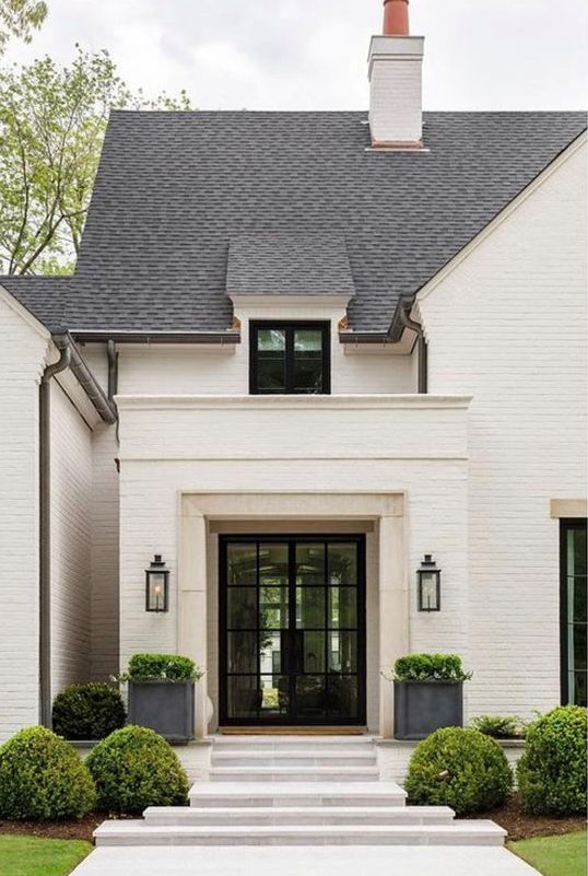 a beautiful modern entrance with a glass door, potted topiaries and some more topiaries on the lawn is a super chic space