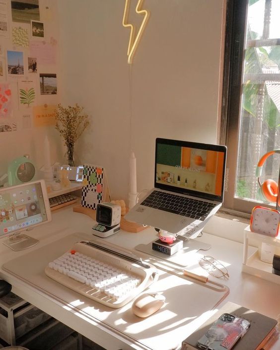 a beautiful neutral workspace with a laptop on a stand, some devices, a gallery wall, some decor on the desk, a neon light and a clock