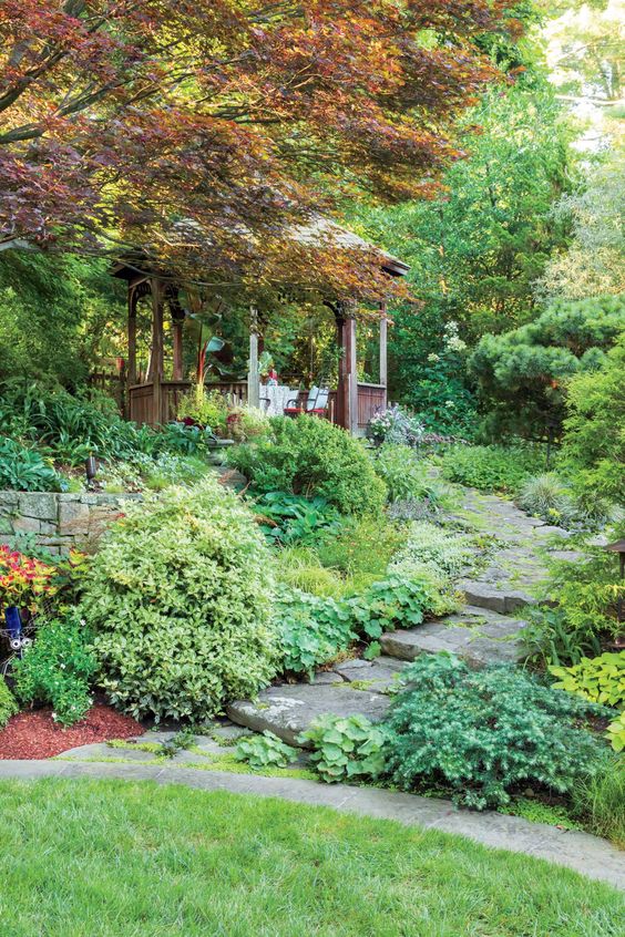 a beautiful shady garden with a grene lawn, greenery, shrubs, a wooden gazebo, a bold fall tree, a stone path is wow