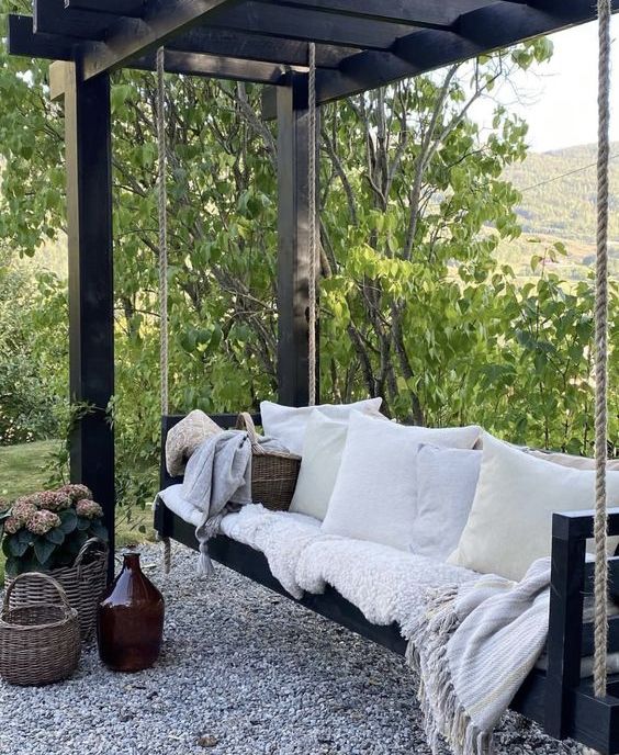 a lovely b&w outdoor space with a dark-stained frame with a swinging daybed and pillows, baskets with blooms is a wonderful space to sleep outdoors and read books