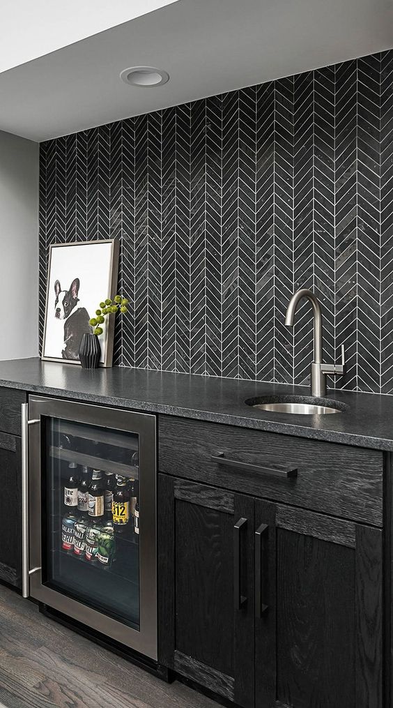 a black kitchen with a black countertop, a black chevron tile backsplash and some pretty modern decor