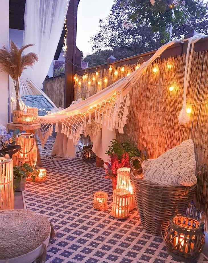 a boho balcony with an embroidered rug, lots of candle lanterns, a hammock with pillows, lamps and pillows and a jute pouf