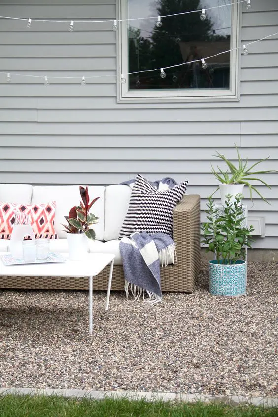 a boho gravel patio with a wicker sofa and pillows, a coffeee table, some potted greenery and lights over the space