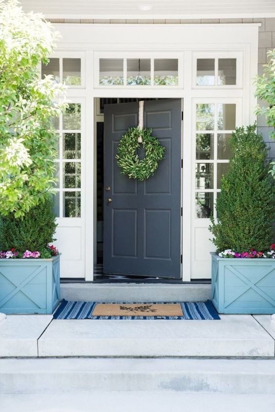 a bold farmhouse porch with a black door, a greenery wreath, layered rugs, potted topiaries and some bold blooms is amazing