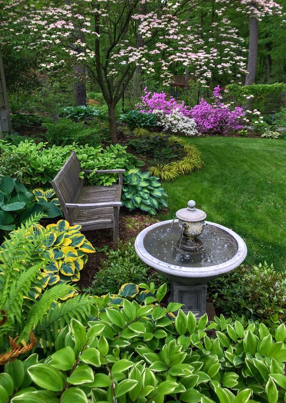 a bold garden with a green lawn, greenery, pink and white blooms, a blooming tree, a bench and a fountain is wow