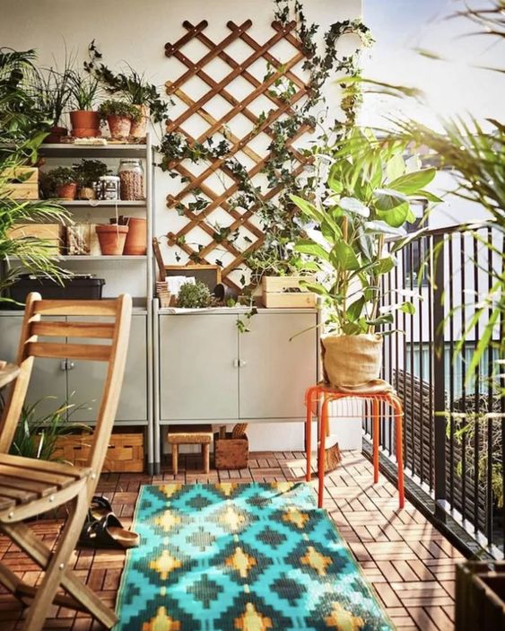 a bright balcony with grey storage and shelving units, a trellis with greenery, potted plants, folding furniture and a bold rug