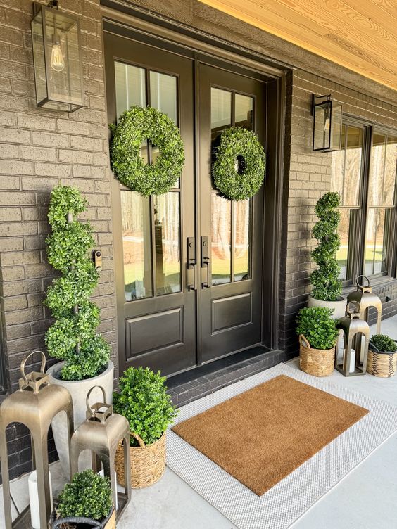 a chic and elegant front porch with black doors, several potted topiaries, candle lanterns and greenery wreaths on the doors