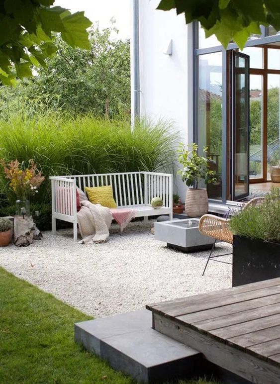 a chic gravel patio with a sofa with pillows, a fire put, a rattan chair, greenery around and some potted plants