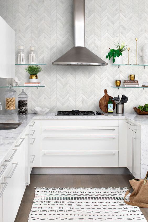 a chic white kitchen with a white marble chevron backsplash, white marble countertops and glass shelves