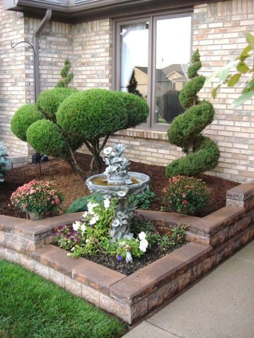 A classic raised bed with stone border is divided into parts and features creatively shaped trees and bushes, a fountain and some blooms. It's a great idea for a refined house entrance.