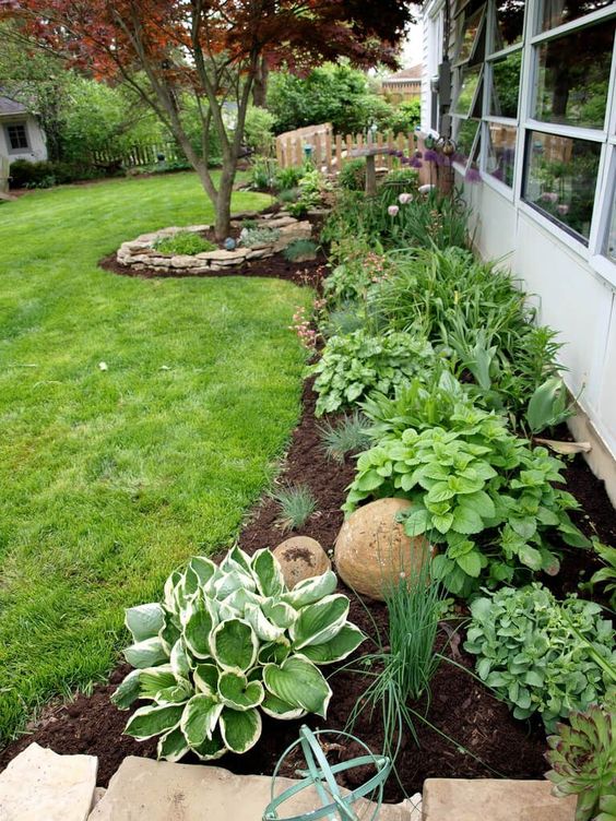 A long garden bed along the house is done with greenery and shrubs, a large stone and a tree will refresh the front yard and all the other outdoor spaces around the house. 