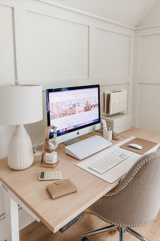 a clean minimal workspace with a desk and a grey chair, some devices and a table lamp and a stand with magazines