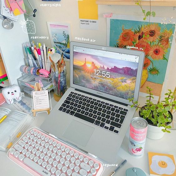 a colorful workspace with a laptop and a pink keyboard, some art, a stand with pens and pencils and pretty decor