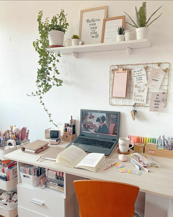 a comfy workspace with a laptop, a stand with pens and pencils, a shelf with artwork and potted greenery and some books