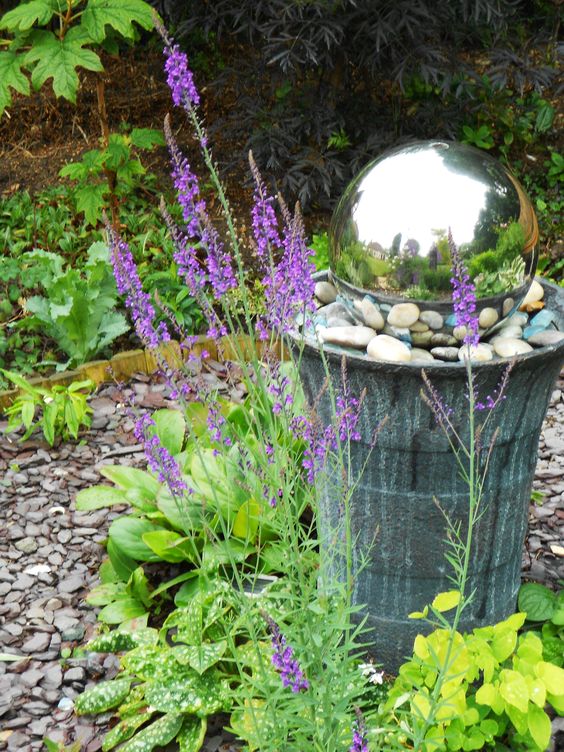 a concrete stand with pebbles and a mirror gazing ball surrounded with blooms and greenery is a bold decoration for a fairy-tale garden