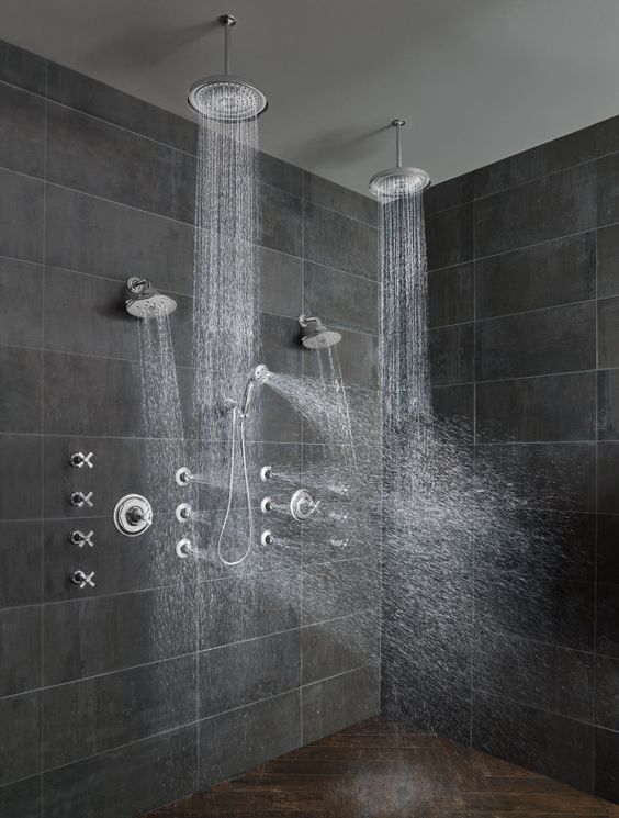 a contemporary shower space with large scale grey stone tiles, a dark stone chevron floor, rain shower heads on the ceiling and some on the wall