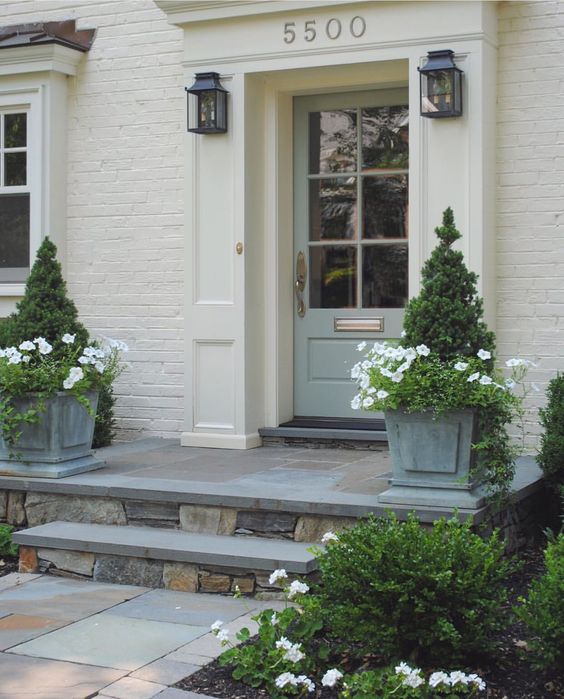 a cool and chic front porch with a glass pane door, two topiaries lining up the entrance and some blooms in the same planters