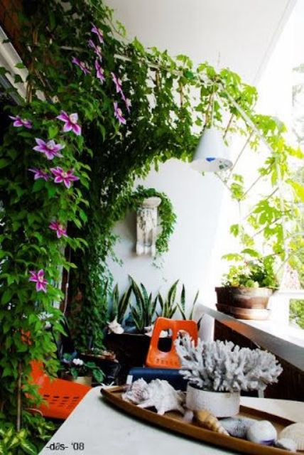 a cool modern balcony with an arched trellis, greenery and blooms coverign it, a table and orange chairs plus some seaside decor