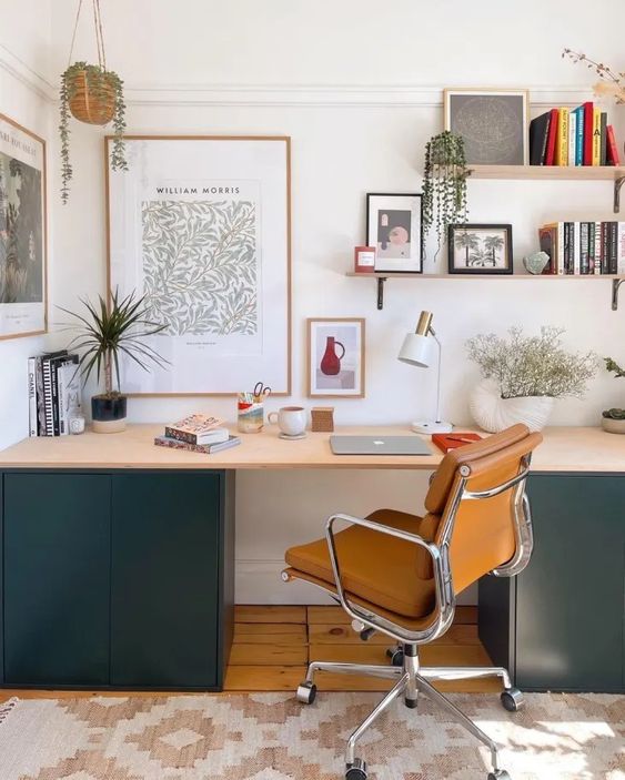 a cool modern workspace with a desk with green cabinets, an amber chair, shelves with books and decor, artworks and a table lamp