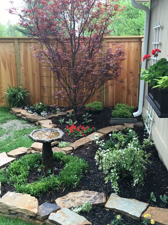 a corner garden with a tree and a raised flower bed with greenery and blooms plus a bird bath is a lovely and cozy space