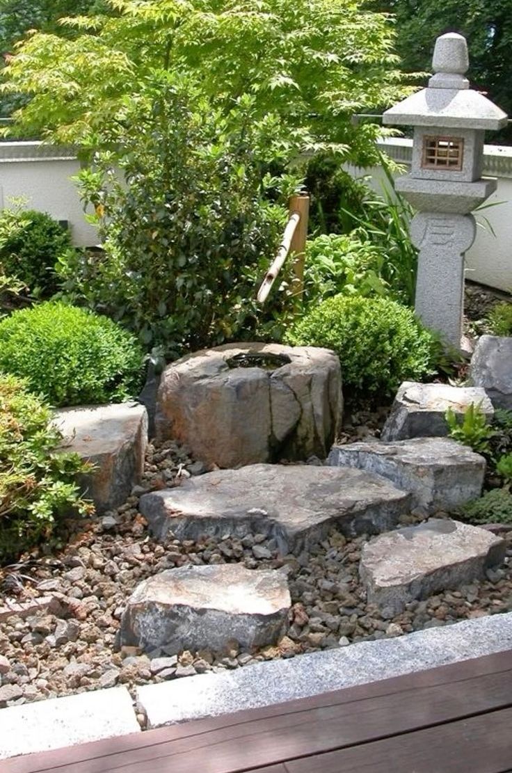 a corner Japanese garden with pebbles and rocks, greenery and topiaries and a tree plus a stone lantern and a bamboo fountain