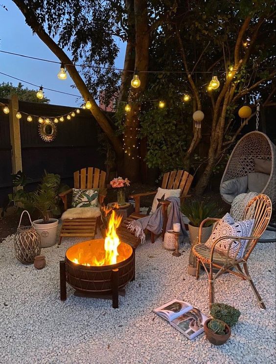 a cozy gravel patio with a fire pit, wooden chairs and a rattan one, potted plants and some blankets and pillows