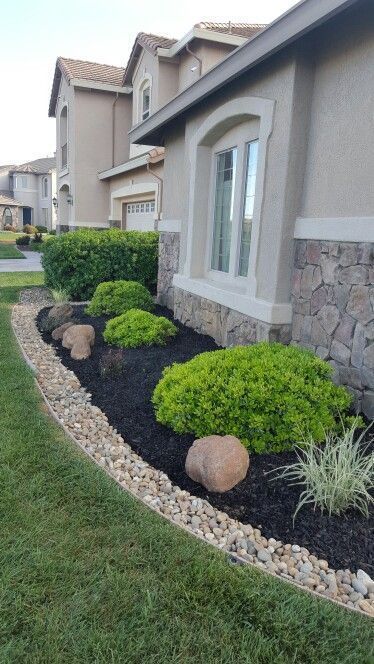 A raised bed covered with black gravel, pebbles, with large stones and greenery is placed along the wall of the house. 