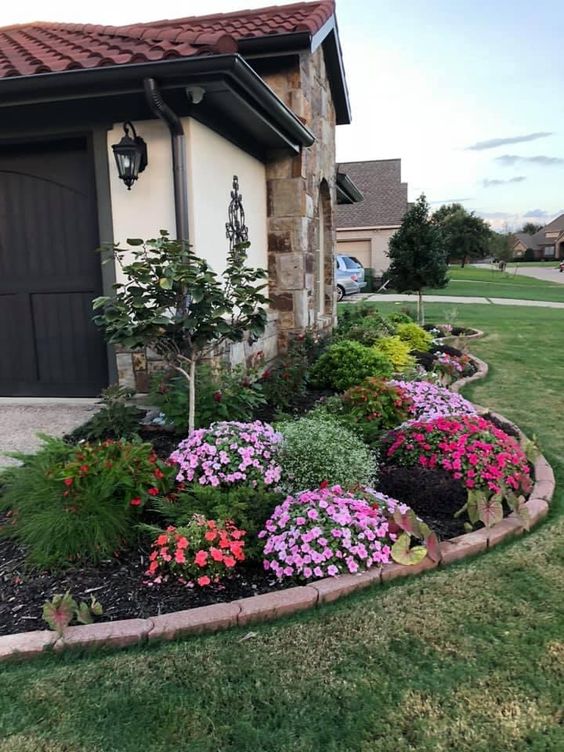 A long curved garden bed with a stone border, with topiaries, a tree and super bright blooms is a catchy decoration for the front yard.
