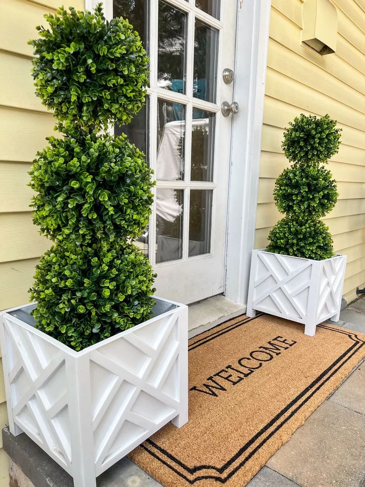 a farmhouse porch with a rug, tall topiaries in white planters and a white glass pane door is a cool space