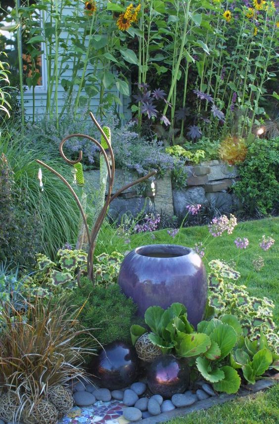 a garden bed with a stone edge, pebbles, a purple vase, bright blooms and greenery and deep purple gazing balls