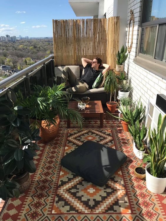 a gorgeous and relaxed boho balcony with a boho rug, a loveseat and a black cushion and lots of cool potted plants around is wow