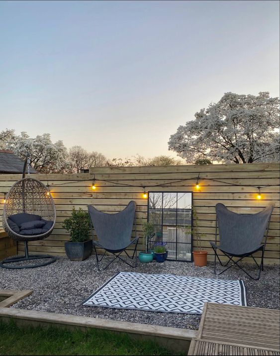 a gravel patio by the fence, grey butterfly chairs, a pendant chair, some potted plants, rugs and lights