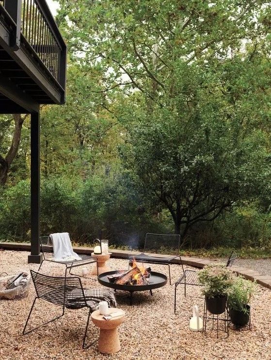 a gravel patio with a black metal fire pit, black metal chairs, a cork side table, potted plants and some lights around