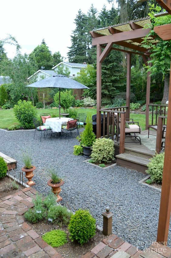 a gravel patio with a dining zone with an umbrella and pillows, a gazebo with forged furniture, some greenery and potted plants around