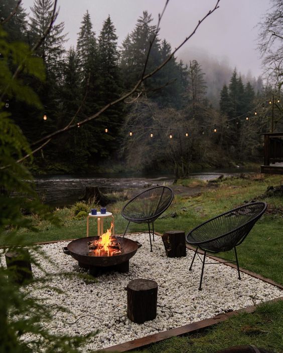 a gravel patio with a fire pit, black chairs, side tables and lights over the space plus a cool river view