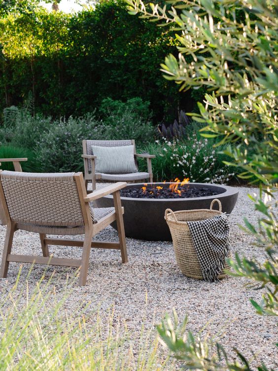 a gravel patio with a fire pit, outdoor chairs, a basket and some greenery and trees around is a cool space to stay