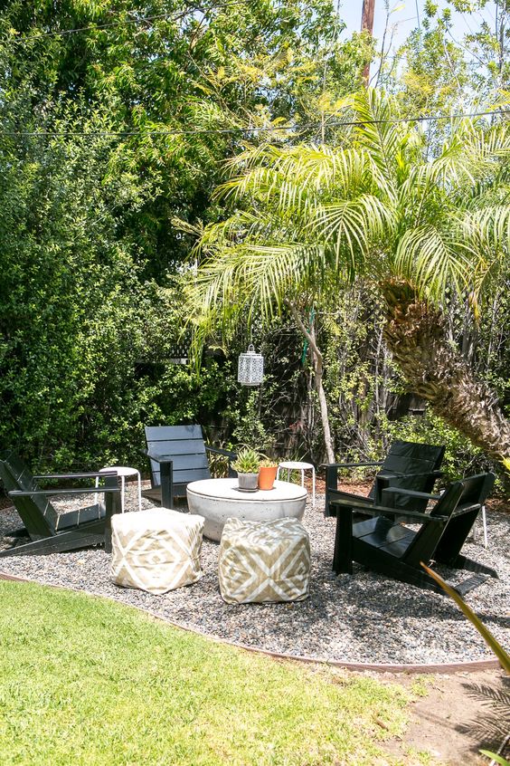 a gravel patio with black wooden chairs, a coffee table, some poufs, lanterns and greenery and trees around