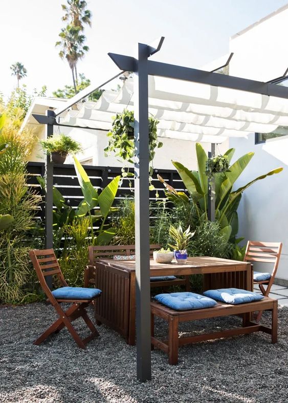 a gravel tropical patio with a black fence, tropical plants, stained dining furniture, blue cushions and a roof