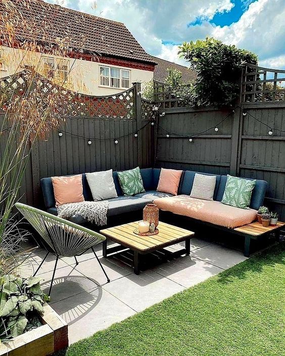 a grey wooden fence with lights, a navy corner sofa with colorful pillows, a coffee table and a round chair plus various plants