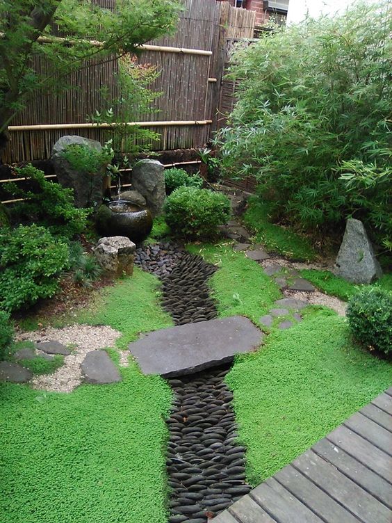 a Japanese-style corner garden with a green lawn, a pebble faux river bed, a classic Japanese fountain, some shrubs and trees around