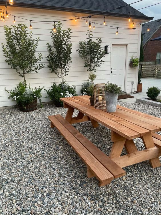 a large farmhouse patio with a wooden table and benches, potted trees and blooms and lights over the space