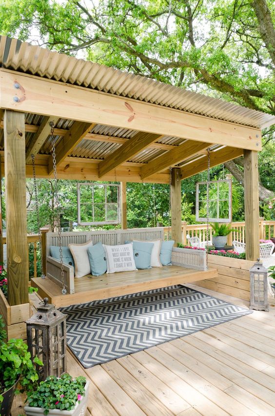 a large swinging bench with pillows placed in a pergola under a roof, surrounded with lanterns and blooms is a cool idea for a rustic or cottage space