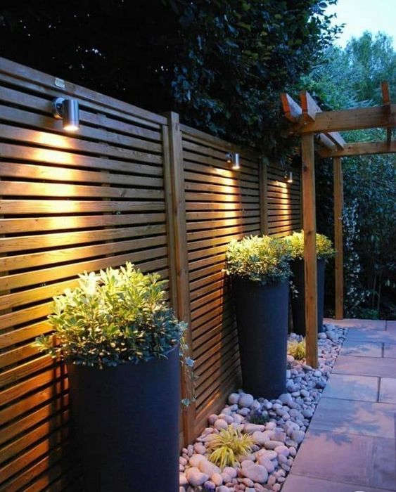 a light-stained planked fence with spotlights, potted greenery in tall planters and pebbles on the ground