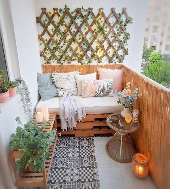 a little cozy balcony with a pallet loveseat and pillows, a planked console table, a side table with blooms and a trellis on the wall with greenery and lights