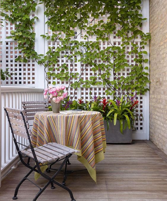 a lovely and cute balcony with a trellis as a privacy screen with greenery, a planter with blooms, folding chairs and a table plus some blooms