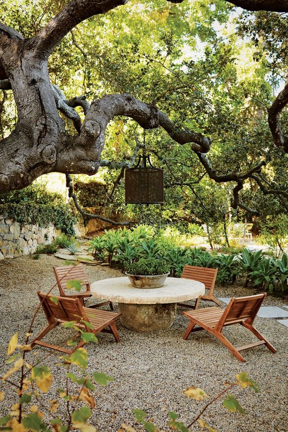 a lovely gravel patio with a stone table, stained chairs, a pendant lamp on the tree and some greenery around the space